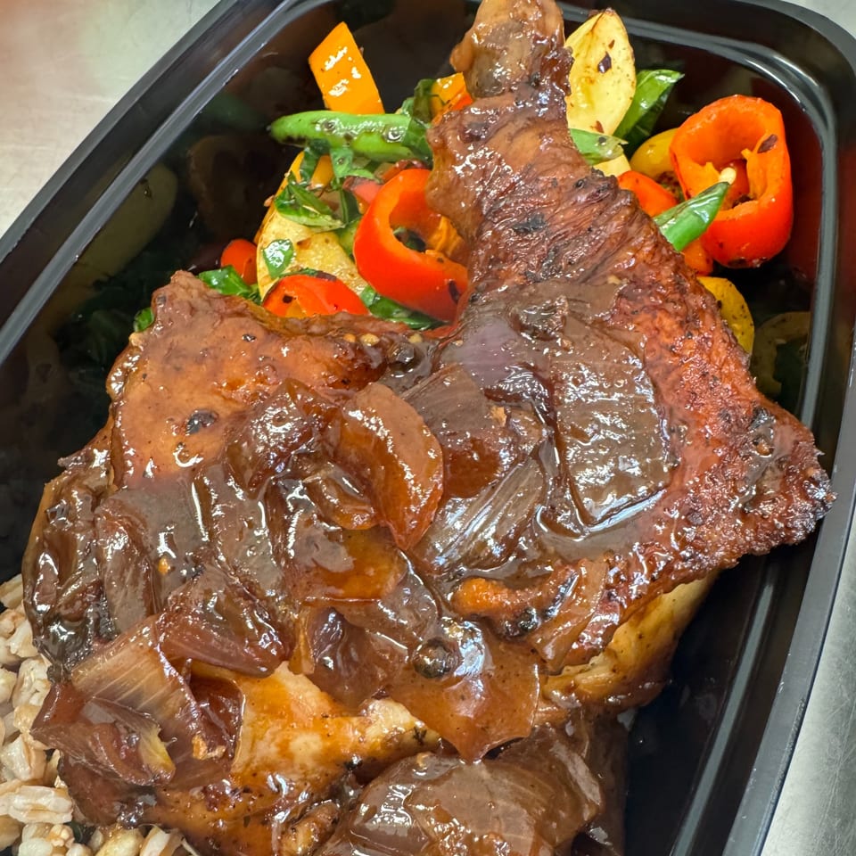 Chicken leg quarter with brown sauce and onions, served with roasted vegetables and grains in a black container.
