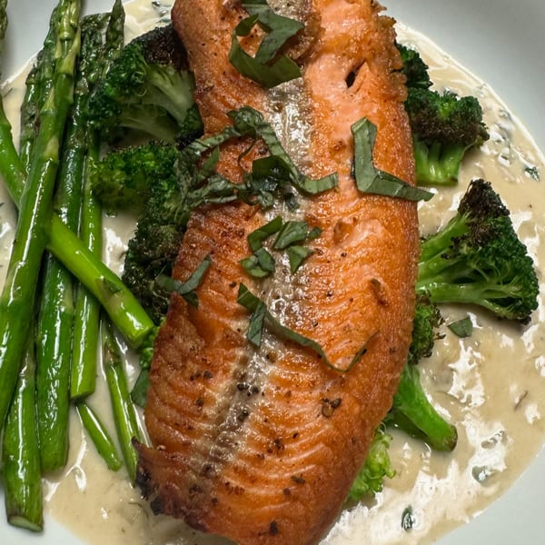 Pan-seared salmon with fresh basil, asparagus, broccoli, and creamy sauce on a white plate.