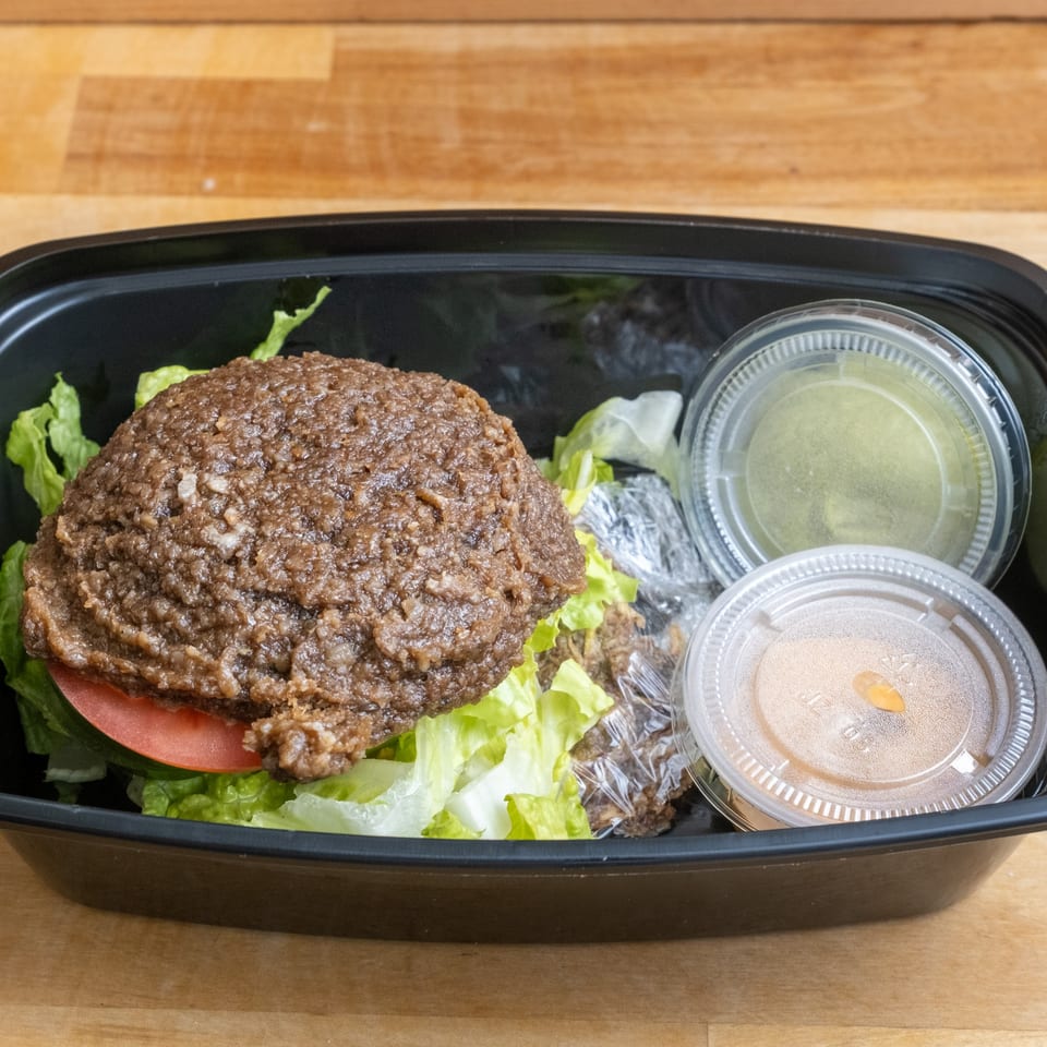 A healthy burger with lettuce and tomato in a black takeout container with two sauce cups.