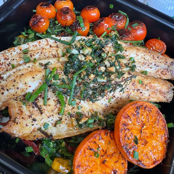 Baked fish fillets with roasted cherry tomatoes, grilled orange slices, and herbs in a black dish.