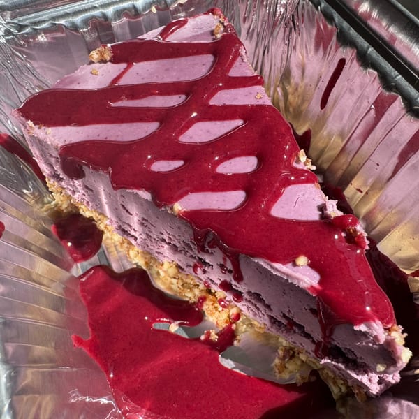 A slice of purple cheesecake with a crumbly crust, drizzled with bright red berry sauce in a foil container.