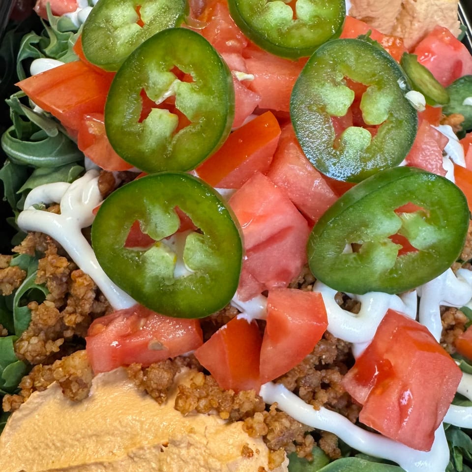 Close-up of a taco salad with ground meat, hummus, tomatoes, jalapeños, and white sauce.